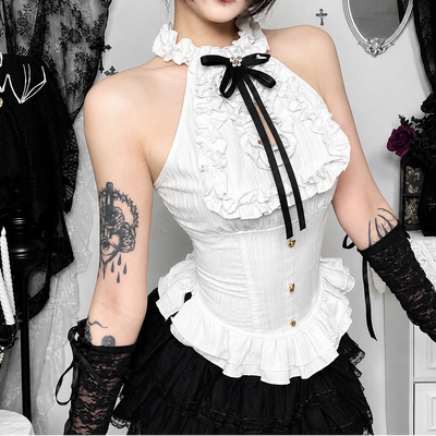 A person wearing a white ruffled halter-style corset top with black ribbon accents, paired with a black ruffled skirt and black lace gloves, photographed indoors against a light wall with clothing racks and decor in the background.