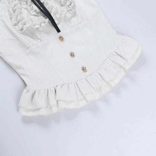 A close-up of a white ruffled top or dress bodice with a double ruffle neckline, three decorative gold-tone buttons, and a black ribbon detail, laid flat on a white background.
